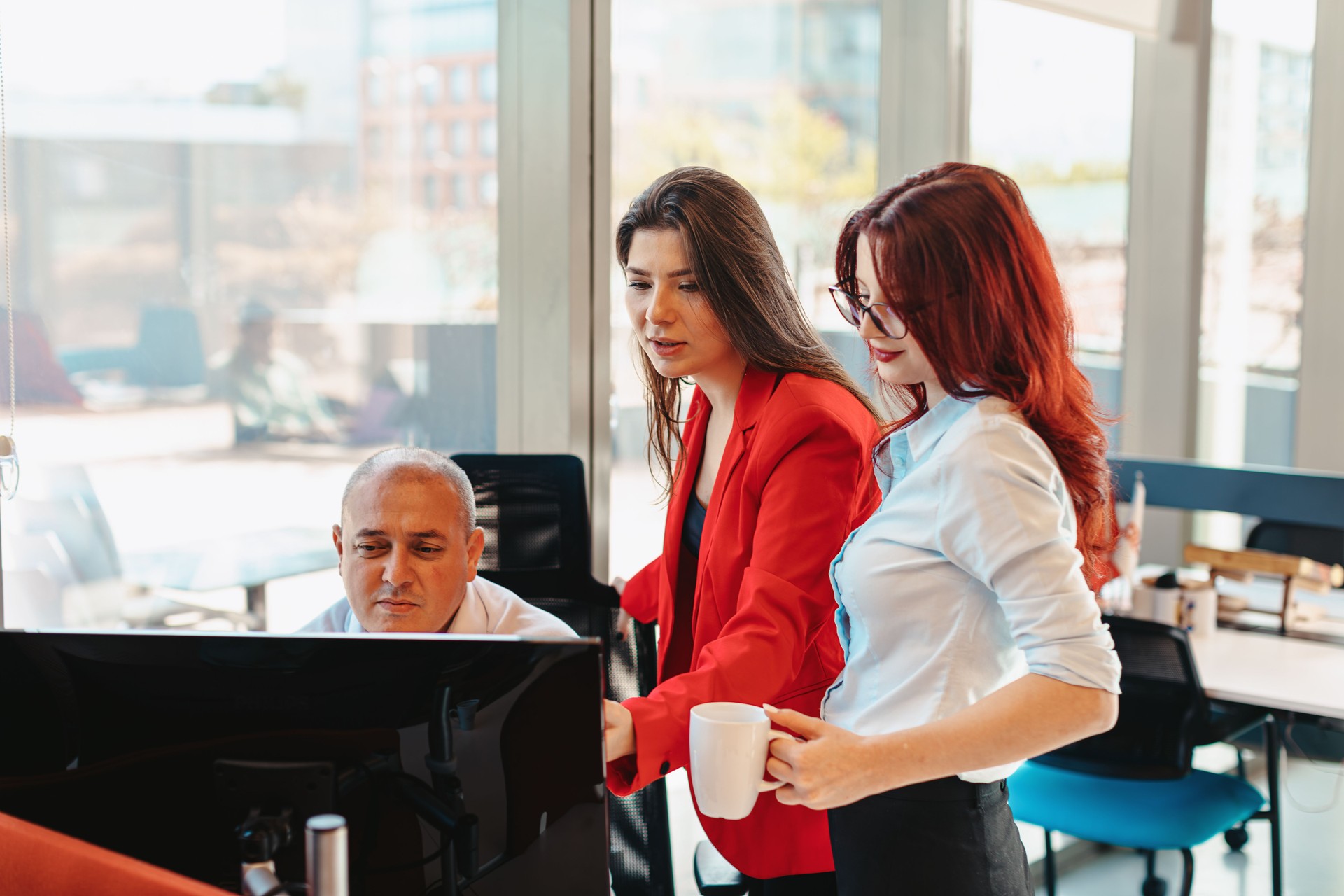 Positive Teamwork in the Office: Young two businesswomen standing by mature businessman working at his desk. Two business partners discussing work in office.